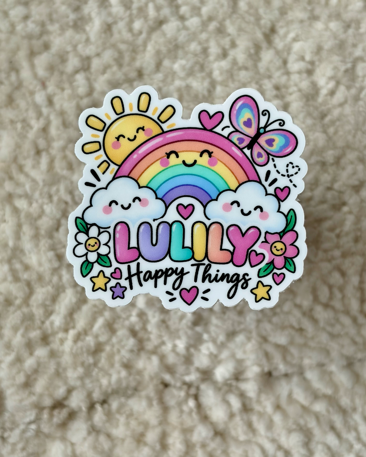A die-cut vinyl sticker featuring a colorful cartoon design with a rainbow, smiling sun, clouds, flowers, butterflies, and the text 'LULILY Happy Things', placed on a beige textured fabric background.