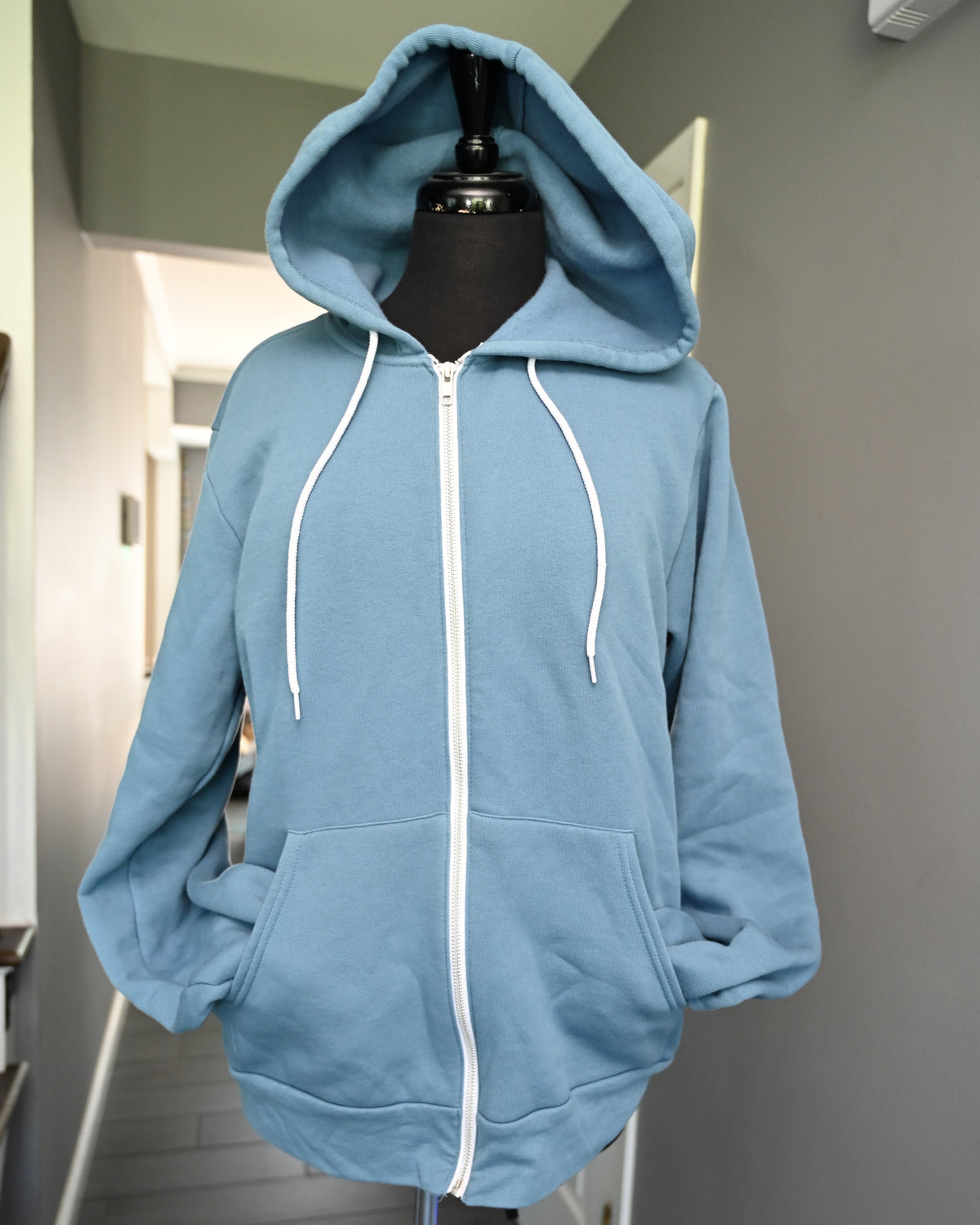 A light blue zip-up hoodie with white drawstrings and a white front zipper, displayed on a black mannequin torso in an indoor hallway.