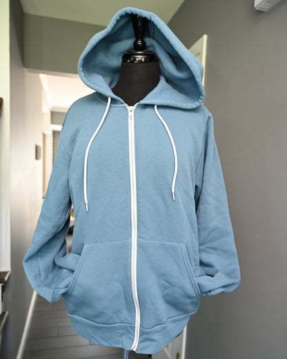 A light blue zip-up hoodie with white drawstrings and a white front zipper, displayed on a black mannequin torso in an indoor hallway.
