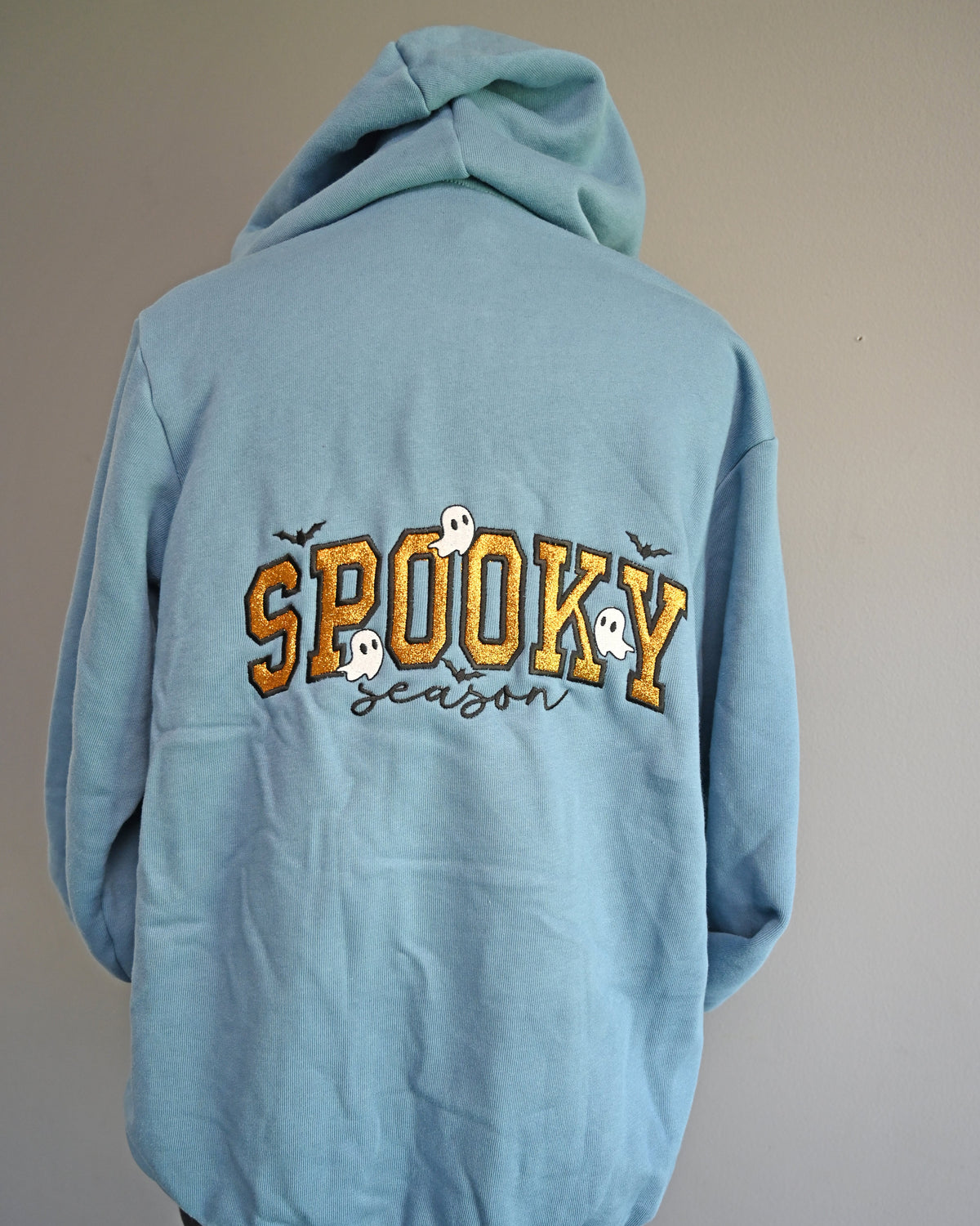 A light blue hoodie shown from the back, displayed on a stand against a plain gray wall. The back features a graphic with the word 'SPOOKY' in yellow outlined letters, decorated with small white ghosts and black bats, above the word 'season' in black script.