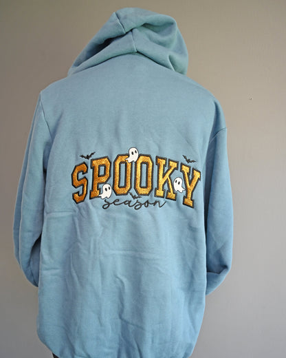 A light blue hoodie shown from the back, displayed on a stand against a plain gray wall. The back features a graphic with the word 'SPOOKY' in yellow outlined letters, decorated with small white ghosts and black bats, above the word 'season' in black script.
