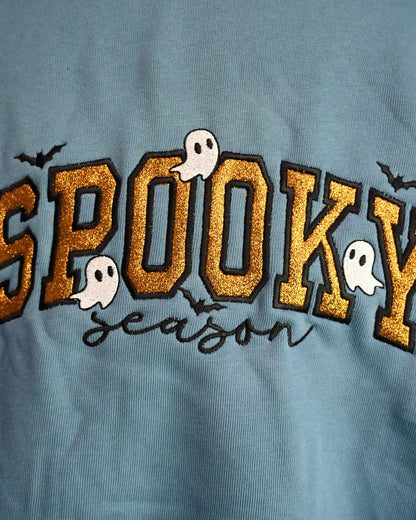 A close-up of a dusty blue sweatshirt featuring a 'Spooky Season' graphic. The main text 'SPOOKY' is embroidered in a glittery gold arched font with black outlines, decorated with small white cartoon ghosts and black bats.