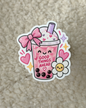 A die-cut sticker featuring a cute pink bubble tea character with a smiling face, pink bow, whipped cream, black pearls, and the text 'GOOD THINGS AHEAD', placed on a beige textured fabric background.