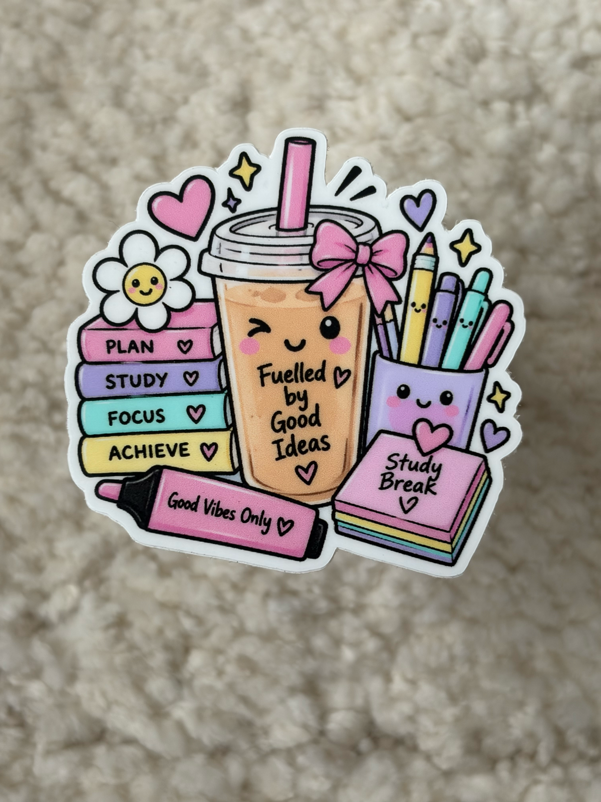 A die-cut sticker featuring a cute cartoon illustration of a pastel drink, books, pens, and motivational words like 'Study Break' and 'Good Vibes Only', placed on a textured beige surface.
