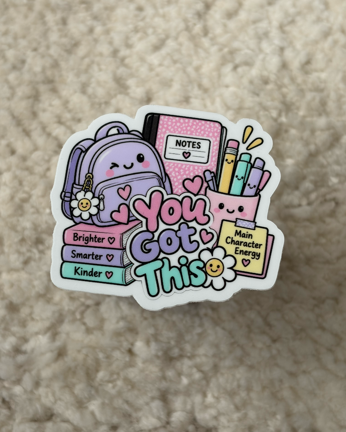 A die-cut sticker featuring a cute school-themed illustration with a purple backpack, books, notebooks, and the words 'You Are Capable' in pastel colors, placed on a beige textured fabric background.
