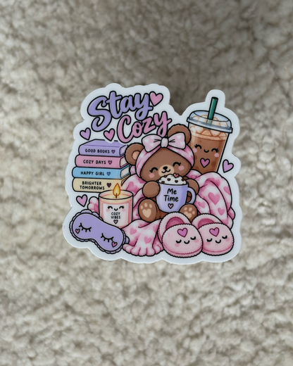 A die-cut sticker featuring a cute cartoon teddy bear wrapped in a pink blanket with a pink drink, candles, and a sleep mask, alongside the text 'Stay Cute' and 'GOOD MAMAS', placed on a textured beige carpet.