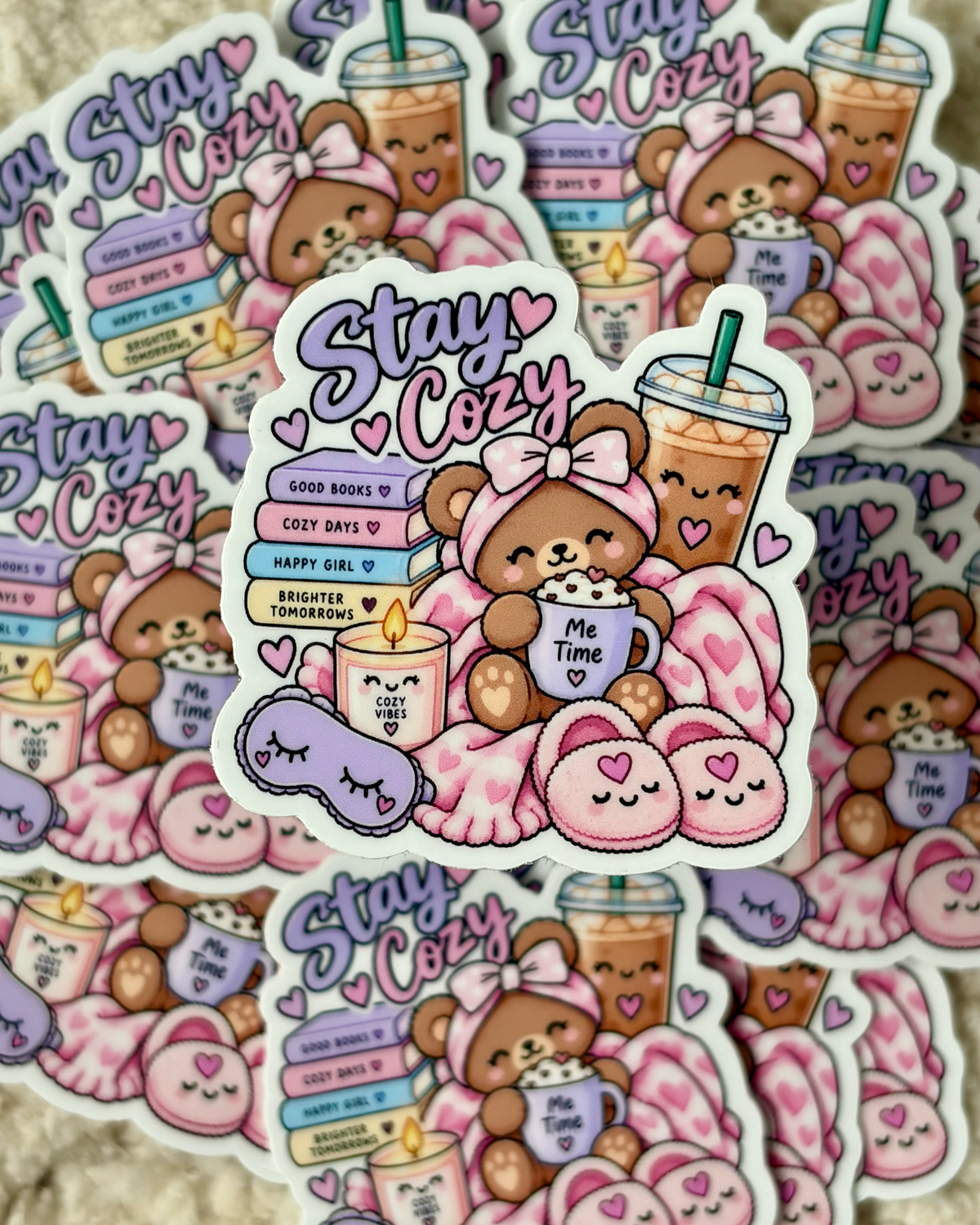 A collection of cute cartoon stickers featuring a cozy teddy bear scene with pink blankets, pink slippers, a purple sleep mask, and iced coffee drinks, all with the text 'Stay Cozy' and hearts, arranged on a soft beige fabric background.