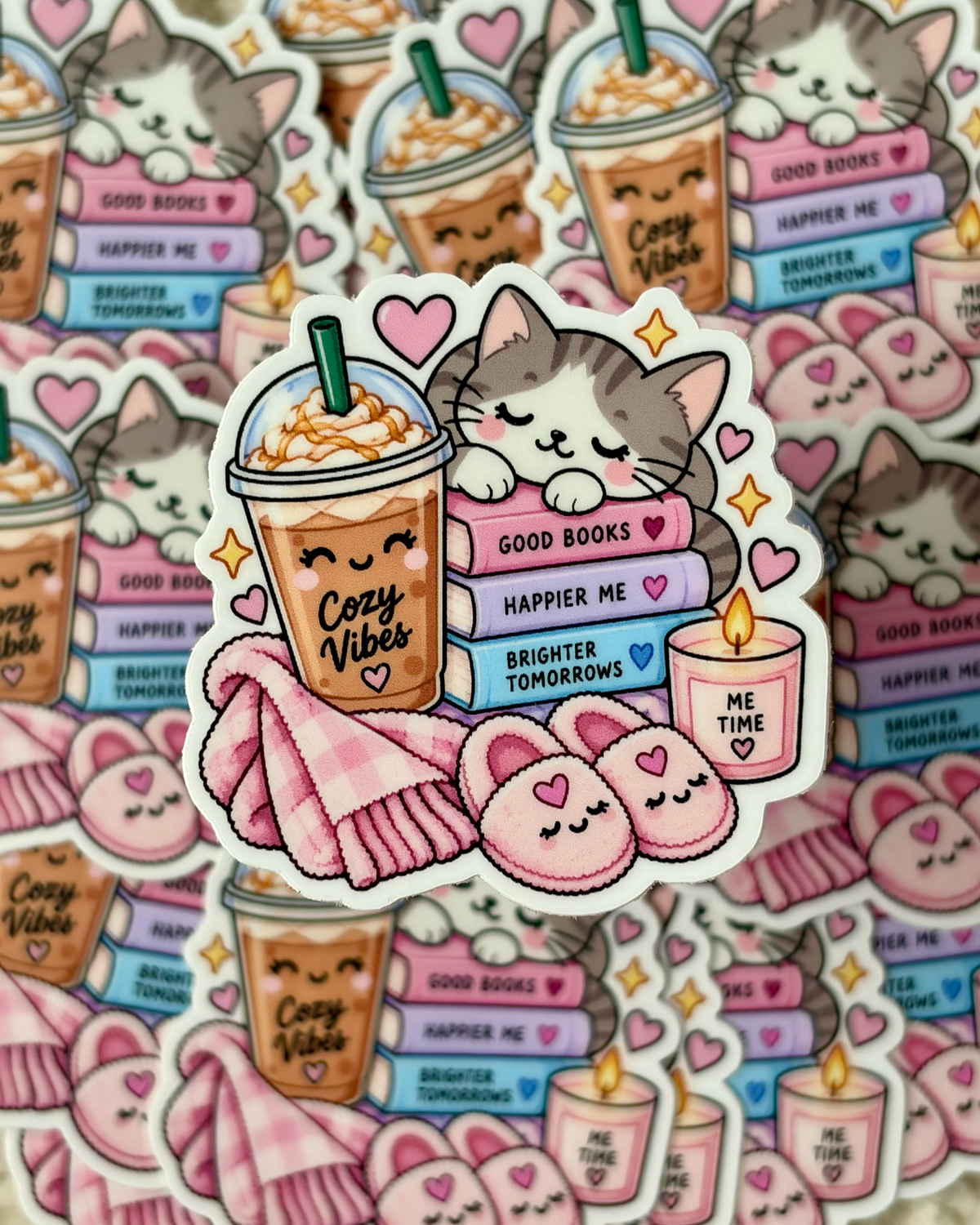 A sheet of cute die-cut stickers featuring a gray and white cat lounging on pink books next to an iced coffee, a pink blanket, slippers, and candles. The center sticker is in sharp focus while matching stickers are scattered in the background.