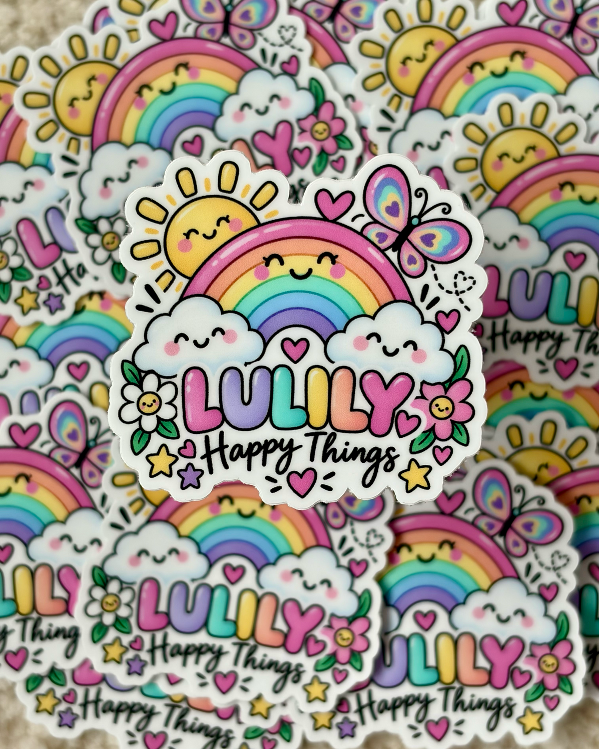 A close-up of a die-cut sticker featuring a smiling rainbow, clouds, flowers, stars, and the text 'LULILY Happy Things,' with multiple identical stickers layered behind it on a soft beige surface.