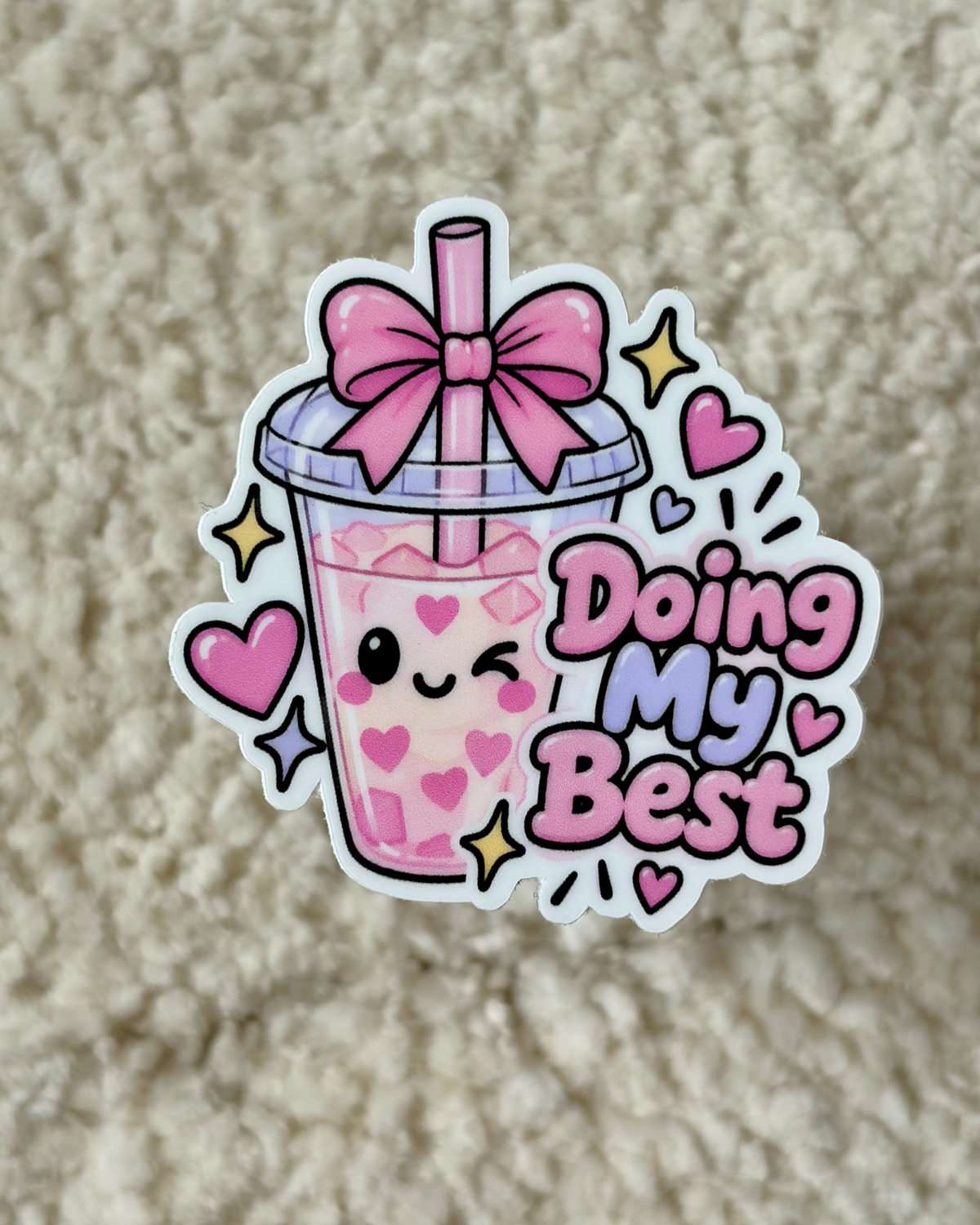 A die-cut sticker featuring a cute pink bubble tea cup with a smiling face, a pink bow on the lid, and the phrase 'Doing My Best' in pink and purple lettering, placed on a beige textured fabric background.