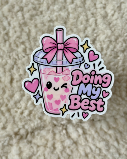 A die-cut sticker featuring a cute pink bubble tea cup with a smiling face, a pink bow on the lid, and the phrase 'Doing My Best' in pink and purple lettering, placed on a beige textured fabric background.