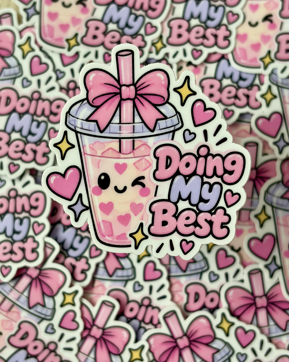 A die-cut sticker featuring a cute pink bubble tea cup with a smiling face, heart details, a pink bow, and the phrase 'Doing My Best' in pink and lavender lettering. The sticker is shown layered over a repeating pile of the same sticker design.