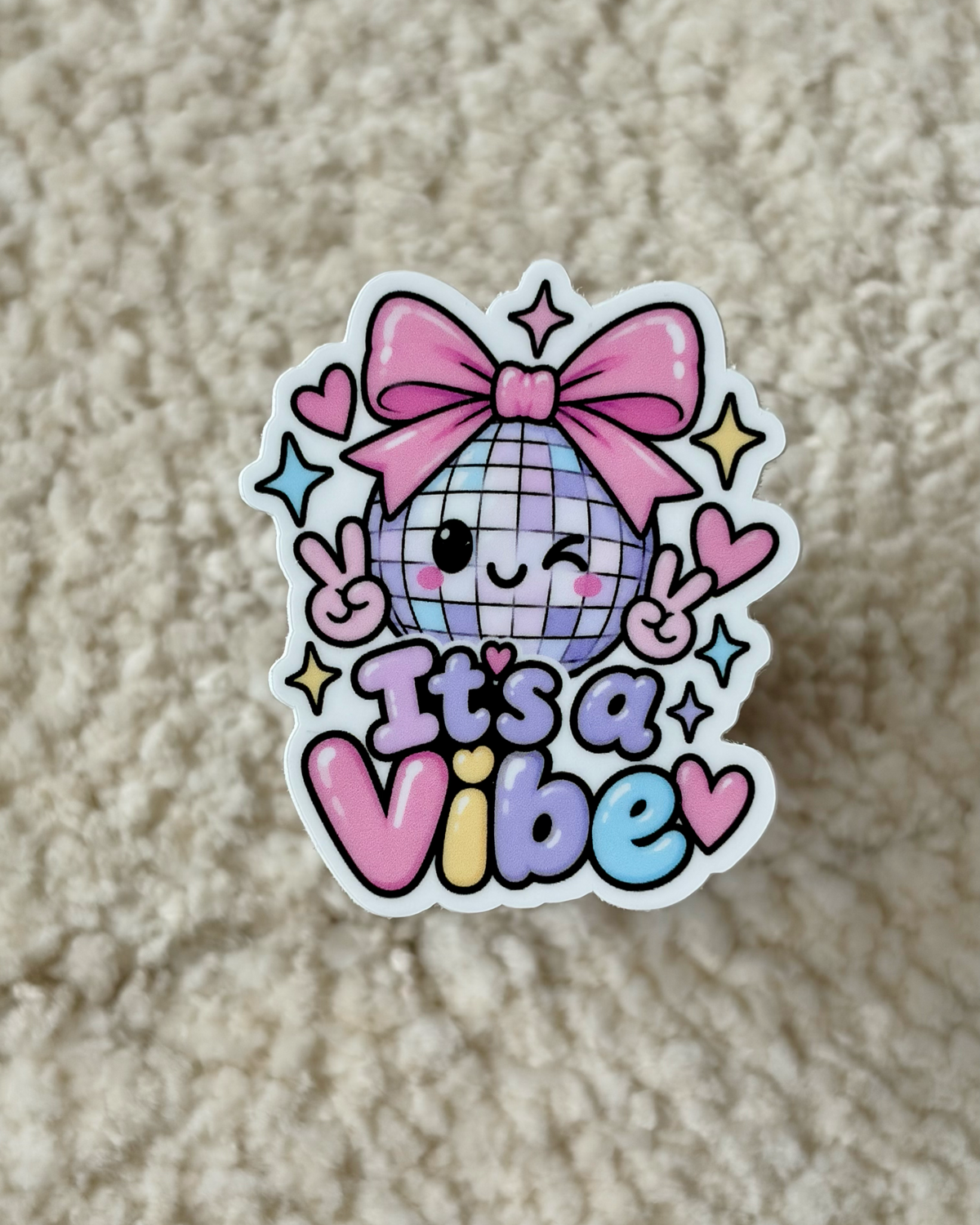 A die-cut sticker featuring a cute disco ball character with a pink bow, winking and making peace signs, with the text 'It's a Vibe' in pastel colors, displayed on a beige textured fabric background.