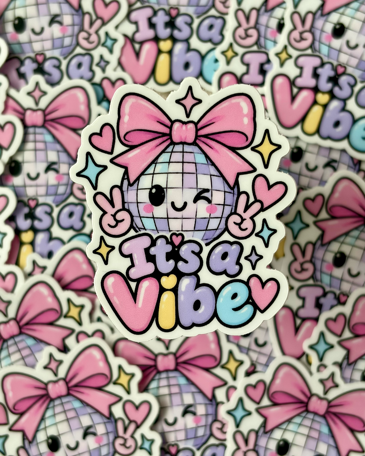 A close-up of a die-cut sticker featuring a cute disco ball character with a pink bow and the text 'It's a Vibe', surrounded by hearts, stars, and peace signs. The sticker is layered over a repeating background of the same sticker design.