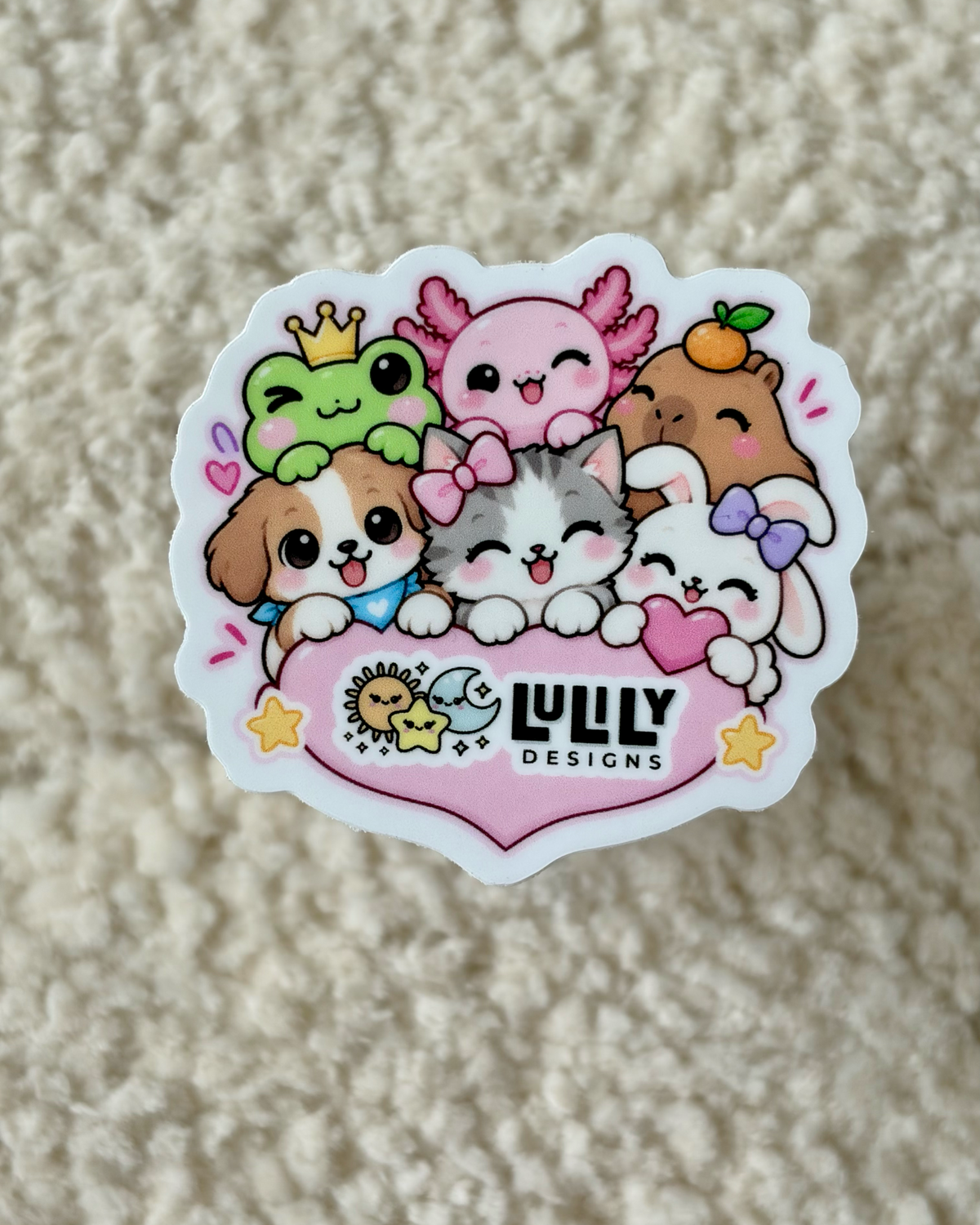A die-cut sticker featuring cute cartoon animals hugging a pink heart with the text 'LULY DESIGNS' in black, placed on a textured beige surface.