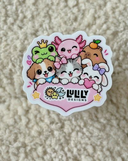 A die-cut sticker featuring cute cartoon animals hugging a pink heart with the text 'LULY DESIGNS' in black, placed on a textured beige surface.