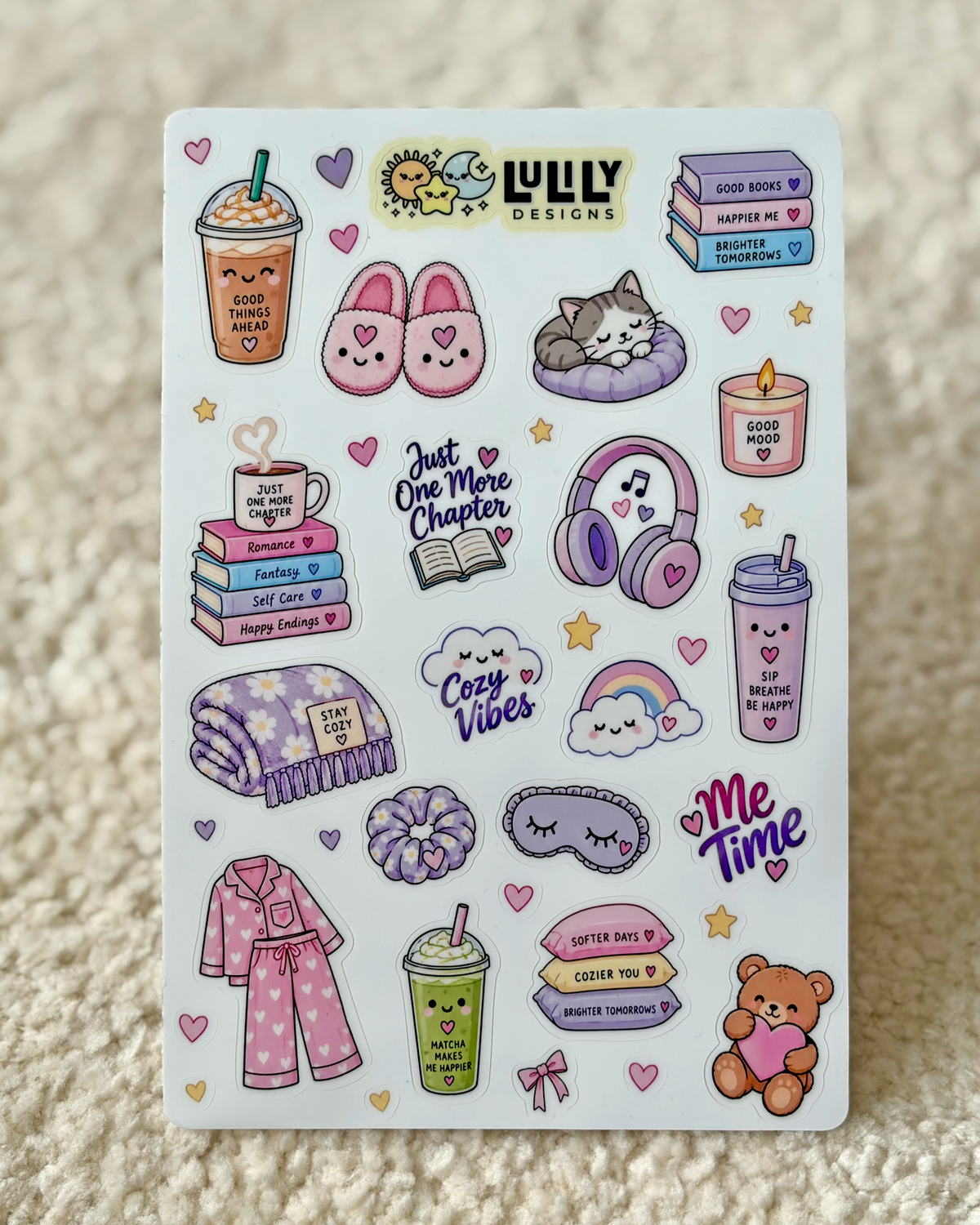 A sheet of cute, cozy planner stickers featuring illustrations of a sleeping cat, pink bunny ears, a cozy drink, books, headphones, pajamas, and a teddy bear, displayed on a textured beige fabric background.