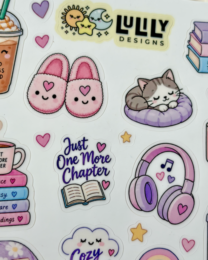 A close-up of a sticker sheet featuring cute pastel illustrations and text stickers, including pink slippers, a sleeping cat on a purple cushion, headphones, books, a coffee drink, stars, hearts, and the brand name LULLY DESIGNS.
