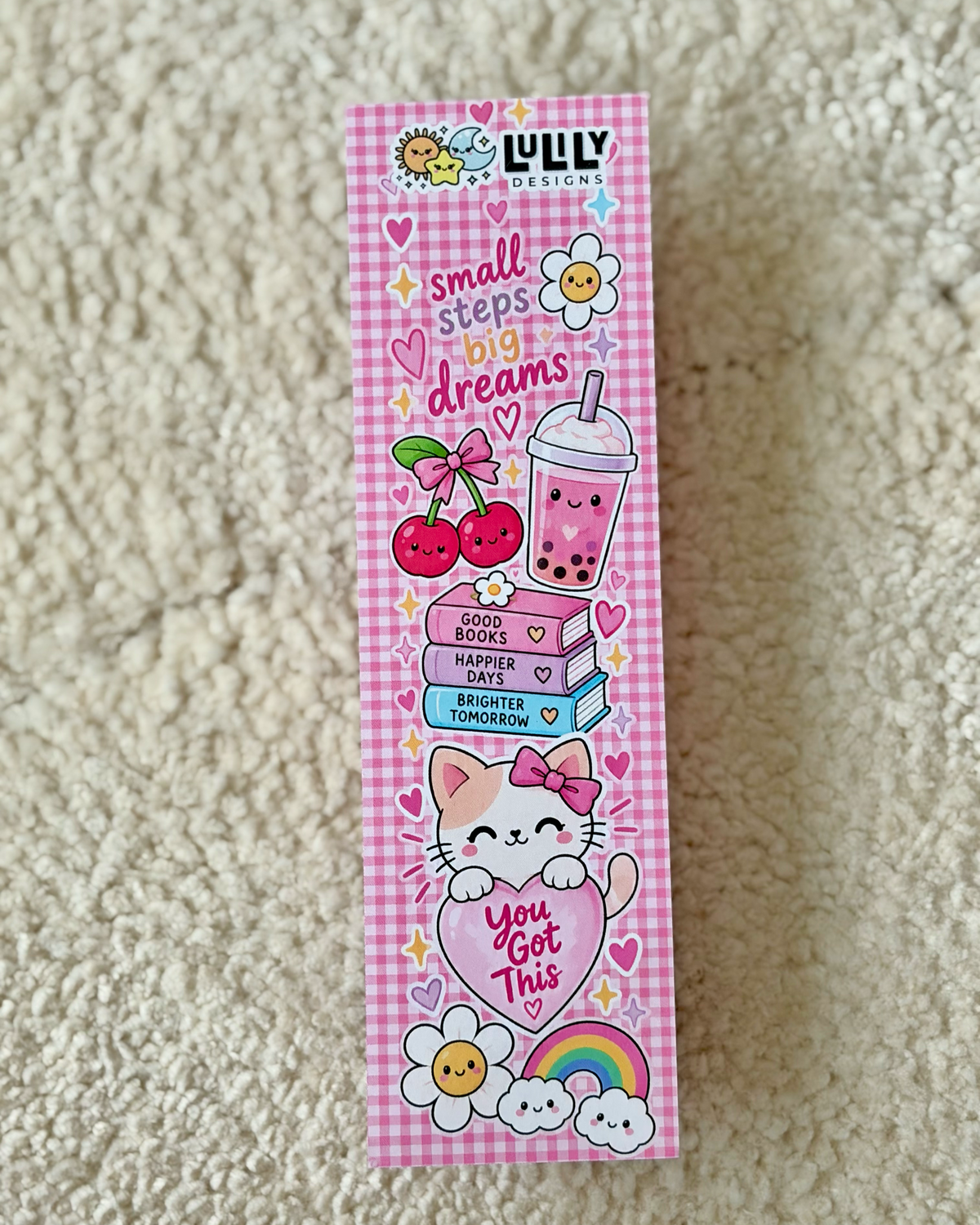 A tall rectangular box for LULY DESIGN stationery, decorated with a pink gingham pattern and cute illustrations including cherries, a drink, books, a cat, flowers, and a rainbow. The box is placed on a beige textured carpet.
