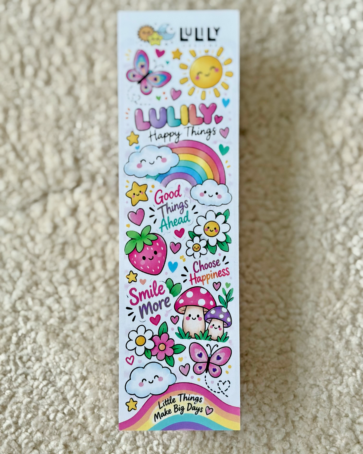 A tall, narrow white sticker sheet featuring colorful cartoon illustrations including a rainbow, butterfly, strawberry, flowers, mushrooms, and text like 'LULILY Happy Things' and 'Good things ahead', placed on a beige textured carpet.