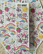 A collection of colorful sticker sheets featuring cute cartoon illustrations including rainbows, butterflies, strawberries, mushrooms, smiling suns, and text like 'LULU Happy Things' and 'Smile More' on a soft beige carpet background.