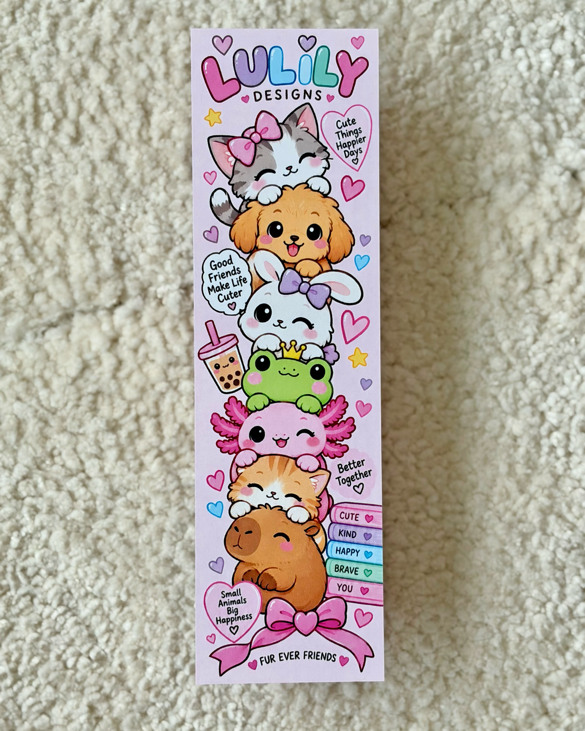 A tall, narrow pink card or sticker sheet featuring cute cartoon animals stacked on top of each other, with the text 'LULILY DESIGNS' at the top and phrases like 'Good friends make life better' and 'Cute together make life happy.' The sheet is photographed lying on a beige textured fabric surface.