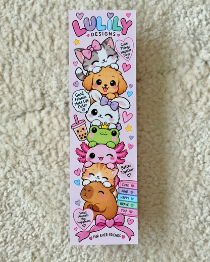 A tall, narrow pink card or sticker sheet featuring cute cartoon animals stacked on top of each other, with the text 'LULILY DESIGNS' at the top and phrases like 'Good friends make life better' and 'Cute together make life happy.' The sheet is photographed lying on a beige textured fabric surface.