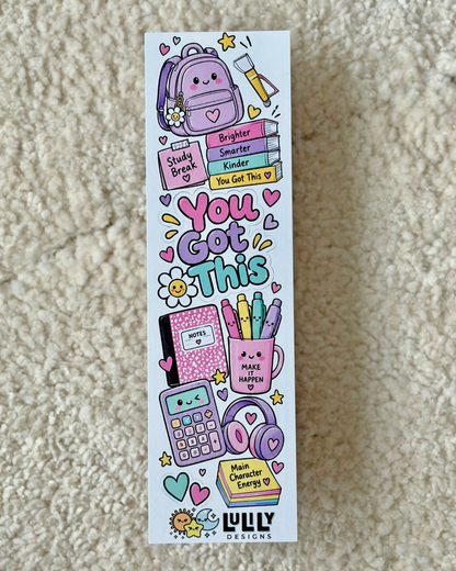 A tall rectangular printed card or sticker sheet with colorful school-themed doodles and motivational text, photographed standing upright on a beige carpet. The design includes a backpack, books, a calculator, headphones, flowers, stars, and phrases like 'You Got This.