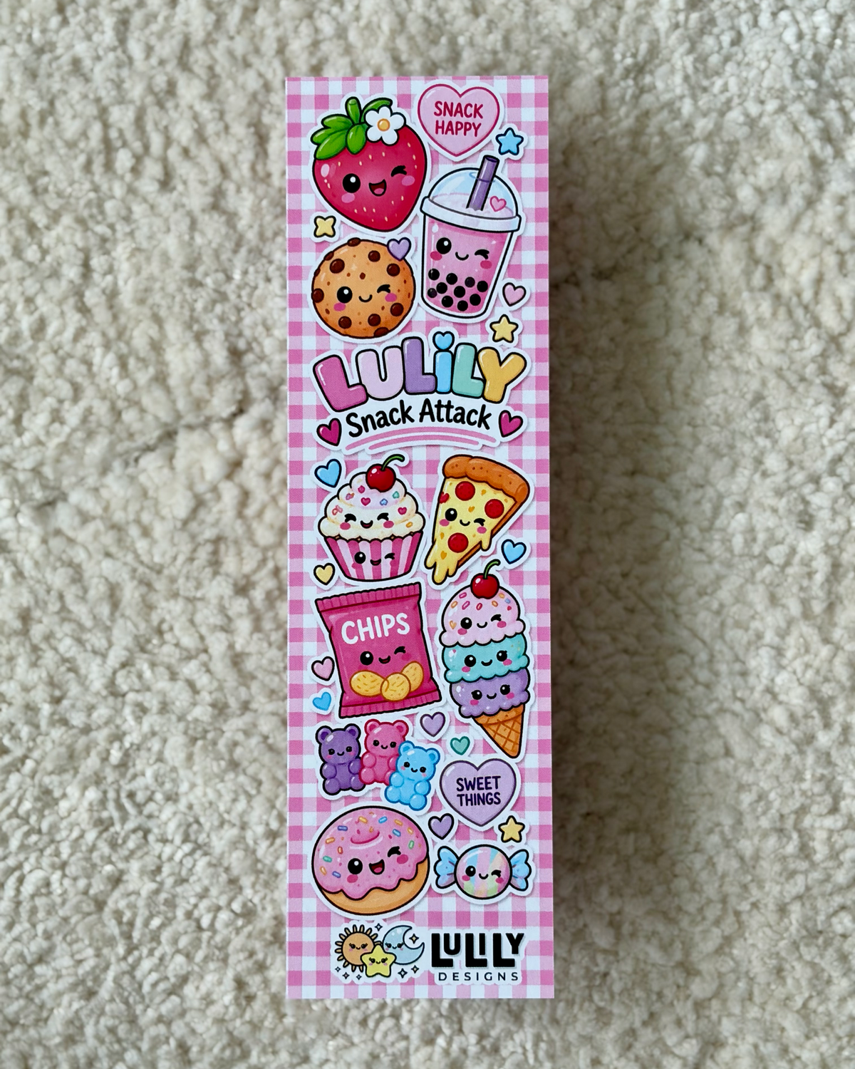 A tall rectangular box for 'Luliy Snack Attack' featuring a pink gingham background and a colorful cartoon illustration of various cute snacks like a strawberry, bubble tea, cookie, cupcake, pizza, ice cream, and donut, placed on a beige textured carpet.