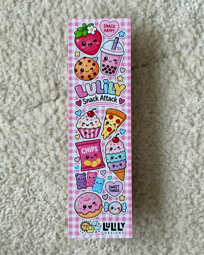 A tall rectangular box for 'Luliy Snack Attack' featuring a pink gingham background and a colorful cartoon illustration of various cute snacks like a strawberry, bubble tea, cookie, cupcake, pizza, ice cream, and donut, placed on a beige textured carpet.