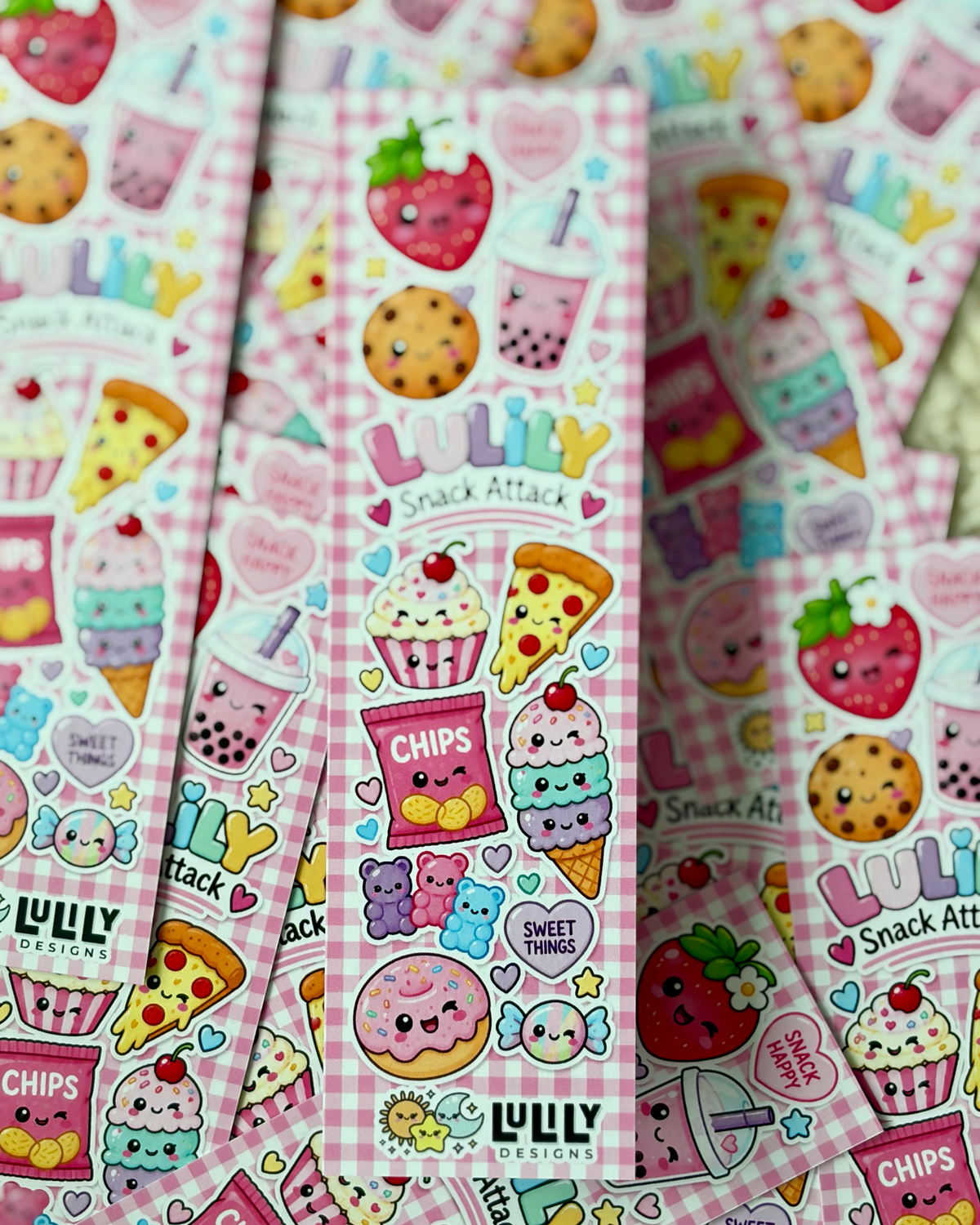 Several narrow pink sticker sheets with a gingham pattern and colorful cartoon snack illustrations are fanned out. The stickers feature kawaii-style strawberries, cupcakes, pizza slices, cookies, boba drinks, donuts, gummy bears, and text including 'LULY DESIGNS' and 'Snack Attack'.
