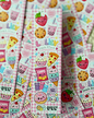 Several narrow pink sticker sheets with a gingham pattern and colorful cartoon snack illustrations are fanned out. The stickers feature kawaii-style strawberries, cupcakes, pizza slices, cookies, boba drinks, donuts, gummy bears, and text including 'LULY DESIGNS' and 'Snack Attack'.
