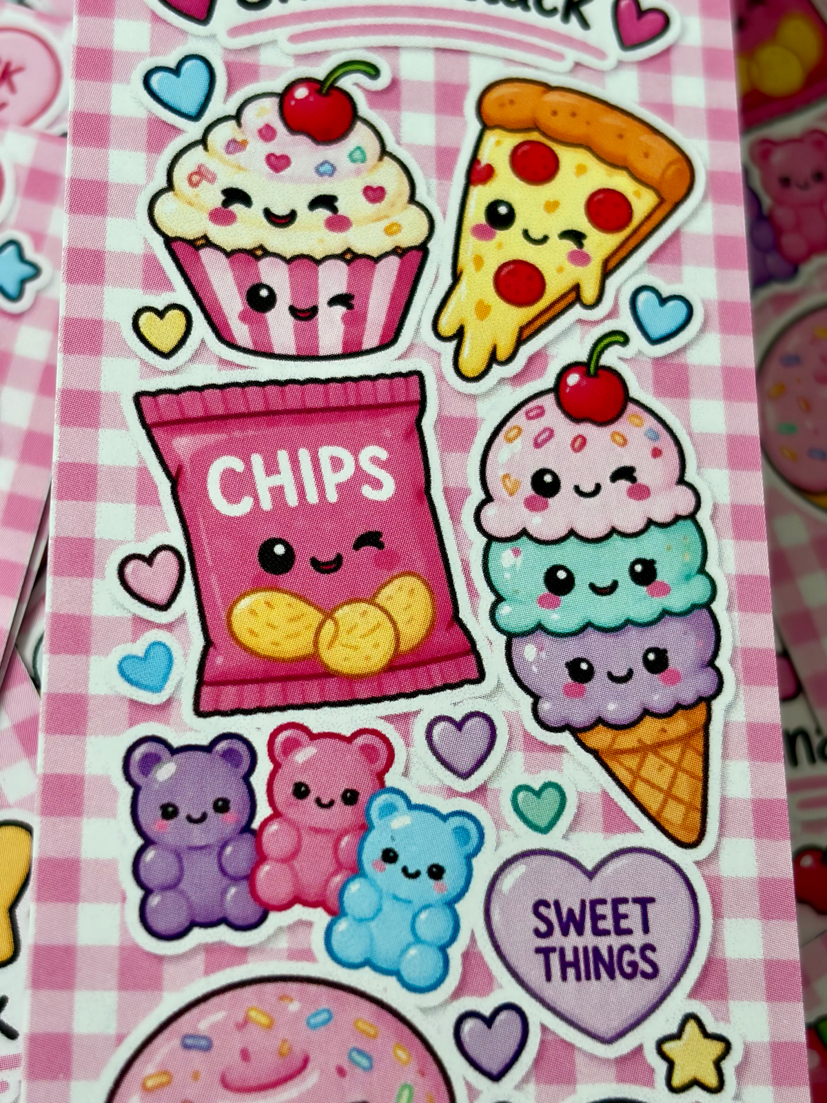 A close-up of a sheet of cute kawaii-style stickers featuring food characters, including a cupcake with a cherry, a pizza slice, a bag of chips, a dessert cone, gummy bears, and hearts on a pink gingham background.