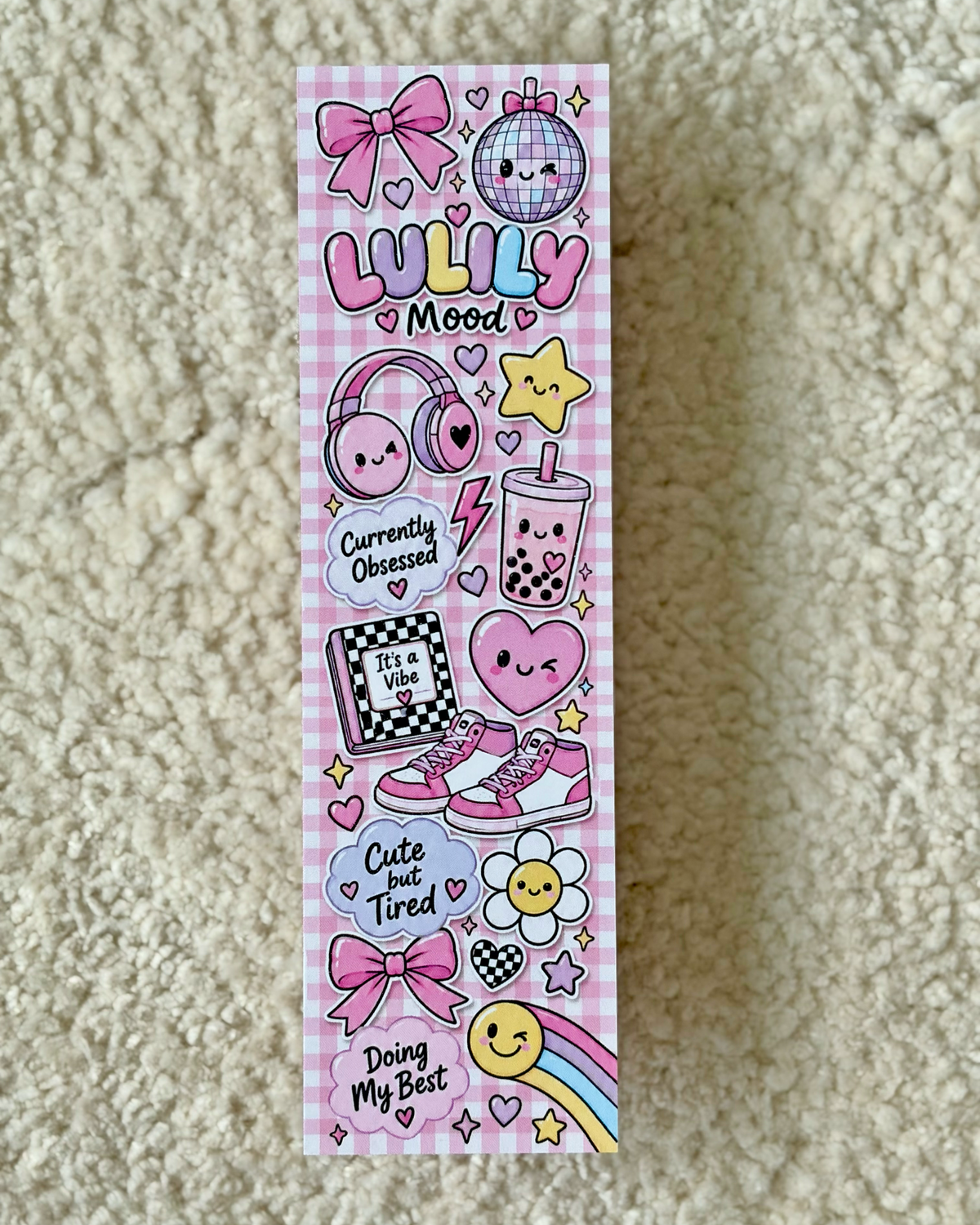 A long rectangular sticker sheet featuring cute kawaii-style illustrations in pink, purple, yellow, and black, including bows, headphones, a disco ball, the words 'LULLY Mood', a boba tea cup, sneakers, flowers, stars, and phrases like 'Currently Obsessed' and 'Cute but Tired', photographed on a beige textured fabric background.