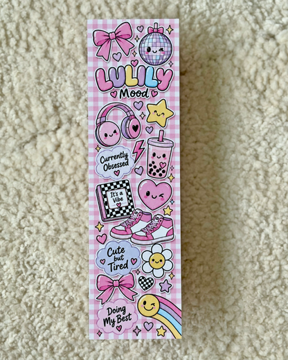 A long rectangular sticker sheet featuring cute kawaii-style illustrations in pink, purple, yellow, and black, including bows, headphones, a disco ball, the words 'LULLY Mood', a boba tea cup, sneakers, flowers, stars, and phrases like 'Currently Obsessed' and 'Cute but Tired', photographed on a beige textured fabric background.