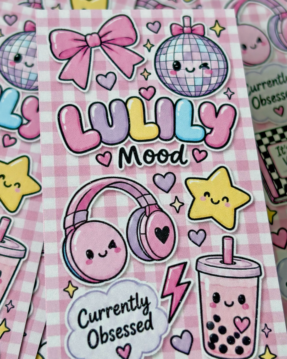 A close-up of a pink gingham sticker sheet featuring cute pastel cartoon graphics, including a disco ball, bow, headphones, stars, a boba cup, and the text 'LULILY Mood' and 'Currently Obsessed'.