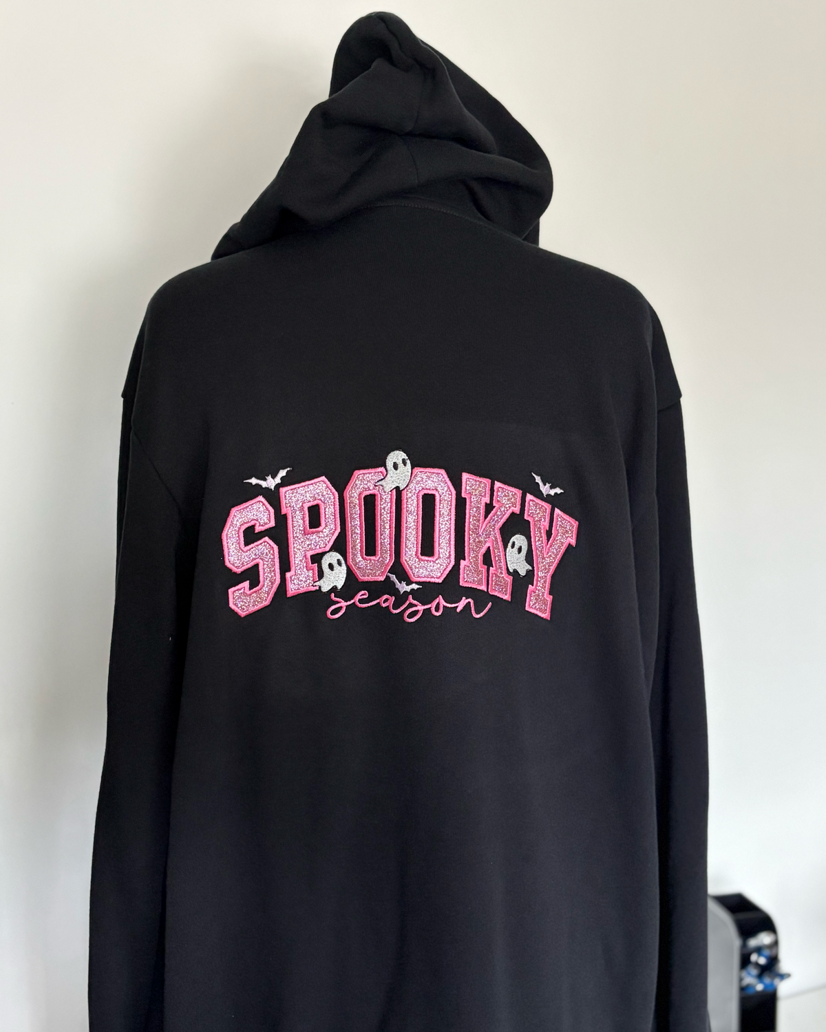 A black pullover hoodie shown from the back, featuring a pink 'SPOOKY season' graphic with small white ghosts and bats on a plain light background.