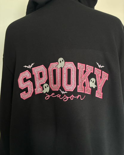 A black hoodie featuring a 'Spooky Season' graphic on the back. The graphic displays 'SPOOKY' in glittery pink varsity-style letters with small white ghosts and bats, and 'season' written in pink cursive underneath.