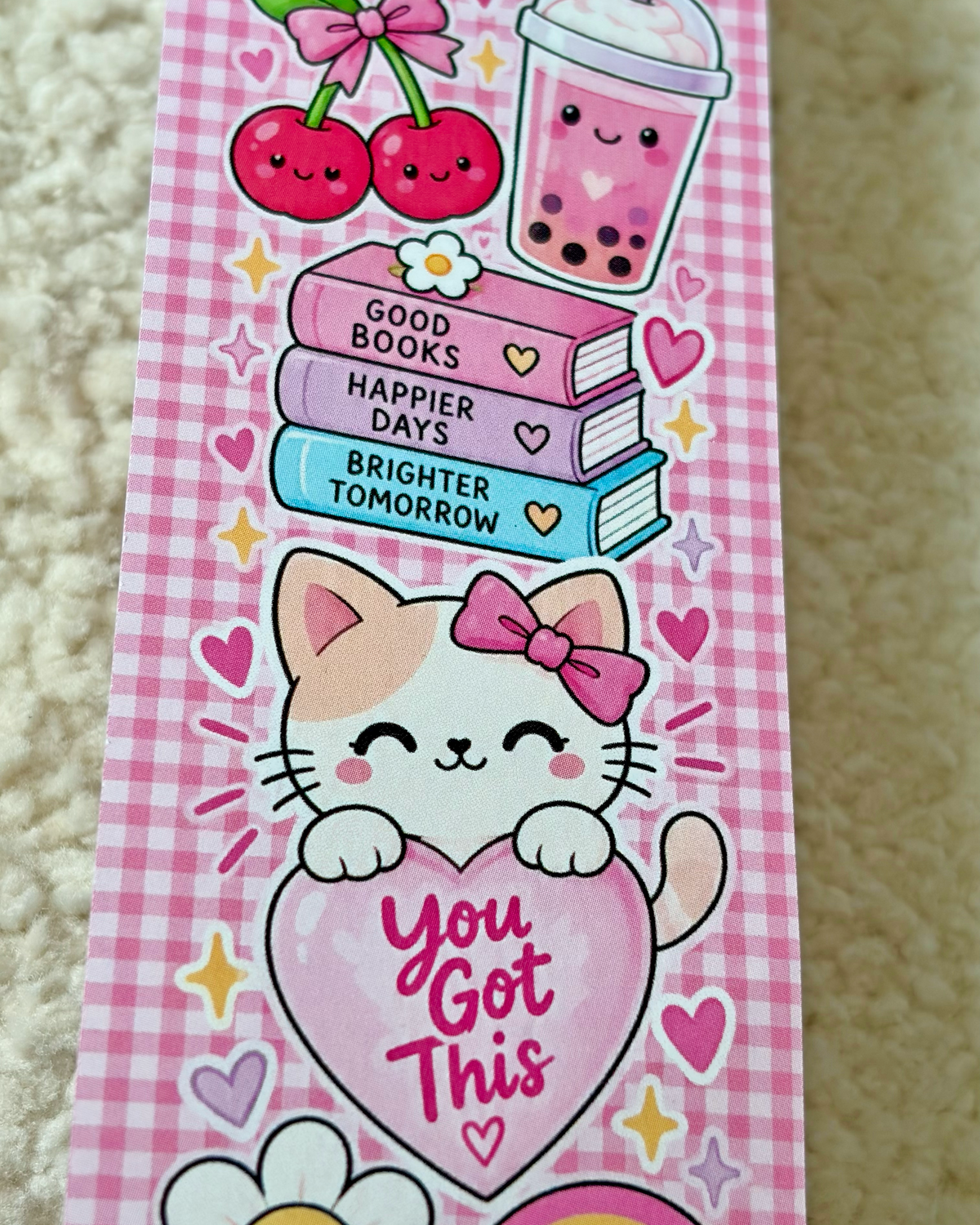 A pink gingham-patterned book marker or bookmark featuring cute cartoon illustrations, including cherries, a bubble tea cup, stacked books with motivational quotes, a smiling cat with a bow, and a large heart that says 'You Got This'.